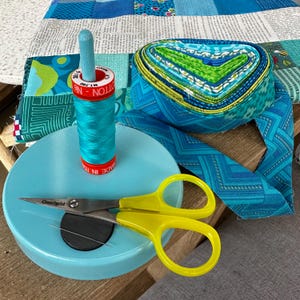 May include: A selection of quilting supplies, featuring a teal thread spool, yellow-handled scissors, and fabric strips in blue and green hues. A blue wooden spool holder and a patchwork quilt are also present.