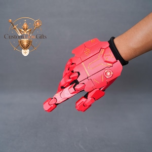Puede incluir: Un guante mecánico articulado de color rojo brillante con placas de armadura segmentadas y detalles de líneas doradas. El guante tiene un puño negro y un diseño complejo y superpuesto que cubre el dorso de la mano y los dedos, creando una estética robótica.