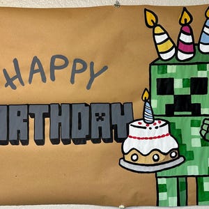 May include: A hand-painted birthday sign featuring a green pixelated character holding a cake with a lit candle. The sign reads "HAPPY BIRTHDAY" in gray block letters on a brown background. Several candles are on the character's head.