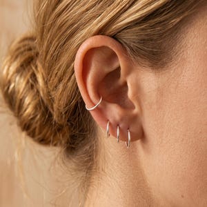 以下が含まれることがあります： 耳に装着された4つの小さく細い銀色のフープピアス。1つは耳の上部の軟骨用カフとして配置され、残りの3つの小さなフープは耳たぶに沿って縦一列に並んでいます。