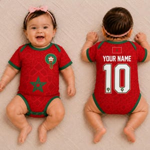 May include: A red baby bodysuit with green trim featuring a Moroccan star emblem on the front and the text YOUR NAME with the number 10 on the back. The garment has a subtle geometric pattern and is worn with a light pink headband.