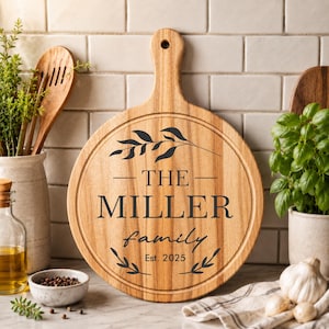 May include: A round wooden cutting board with a handle, engraved with "THE MILLER family Est. 2025" and decorative leaf accents. The board is light brown and appears to be made of wood. It is a kitchen item.
