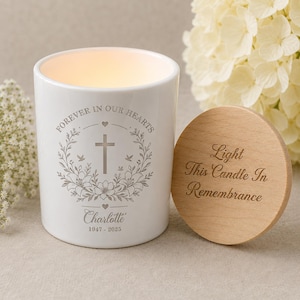 May include: White memorial candle with the text "Forever in Our Hearts" and a cross design. The candle includes the name "Charlotte" and dates 1947-2025. A wooden lid with the text "Light This Candle In Remembrance" is next to the candle.