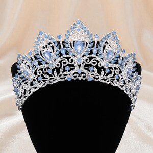 Op de afbeelding: Een zilverkleurige tiara met ingewikkeld metalen filigraanwerk, versierd met talloze lichtblauwe kristallen. Het ontwerp bevat meerdere druppelvormige blauwe stenen die symmetrisch zijn gerangschikt over de gebogen kroonstructuur.