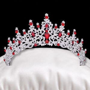 May include: A silver-toned metal tiara featuring an intricate filigree design encrusted with numerous clear white rhinestones and accented with multiple teardrop and round-shaped red crystals arranged in a symmetrical, pointed crown pattern.