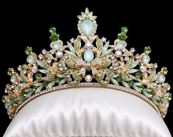 May include: A gold-toned tiara featuring an intricate metal frame adorned with light green and clear crystals, along with oval-shaped opalescent stones. The design includes leaf-like patterns and multiple pointed peaks, creating a symmetrical crown.