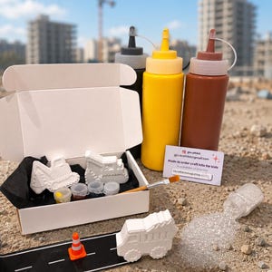 May include: A craft kit featuring a white cardboard box containing two white plaster construction vehicle shapes, four small paint pots, and a paintbrush. Includes three squeeze bottles in black, yellow, and brown, a small orange traffic cone, a black road strip, and a container of silver glitter.