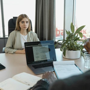 K&ouml;nnte beinhalten: Eine Frau in einem hellgr&uuml;nen Blazer sitzt an einem Tisch gegen&uuml;ber einer Person. Ein Laptop mit einem Dokument, ein Tablet, ein Notizblock und ein Glas Wasser stehen auf dem Tisch. Eine Topfpflanze steht in der N&auml;he eines Fensters.