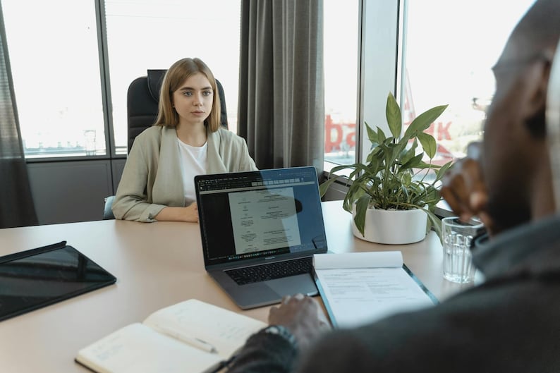 K&ouml;nnte beinhalten: Eine Frau in einem hellgr&uuml;nen Blazer sitzt an einem Tisch gegen&uuml;ber einer Person. Ein Laptop mit einem Dokument, ein Tablet, ein Notizblock und ein Glas Wasser stehen auf dem Tisch. Eine Topfpflanze steht in der N&auml;he eines Fensters.