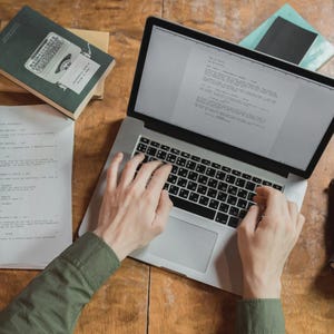 Pu&ograve; includere: Una persona digita su un laptop argentato con tastiera nera, circondata da libri e fogli su una superficie di legno. Lo schermo del laptop mostra del testo. Si vede una camicia verde.