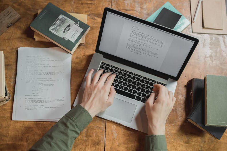 Pu&ograve; includere: Una persona digita su un laptop argentato con tastiera nera, circondata da libri e fogli su una superficie di legno. Lo schermo del laptop mostra del testo. Si vede una camicia verde.