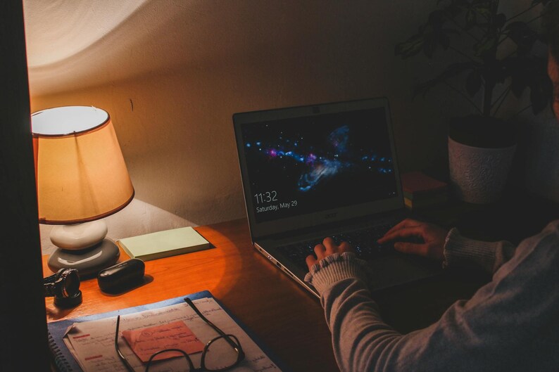 May include: A laptop displaying a colorful galaxy image sits on a wooden desk, with the time 11:32 and the date Saturday, May 25 visible on the screen. A desk lamp with a tan lampshade and a small notepad are also on the desk.