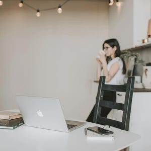 May include: A contemporary workspace with a white table holding a laptop, notebook, and smartphone. A black chair is behind the table. A person is in the background, holding a cup. Overhead, a string of lights adds ambiance.
