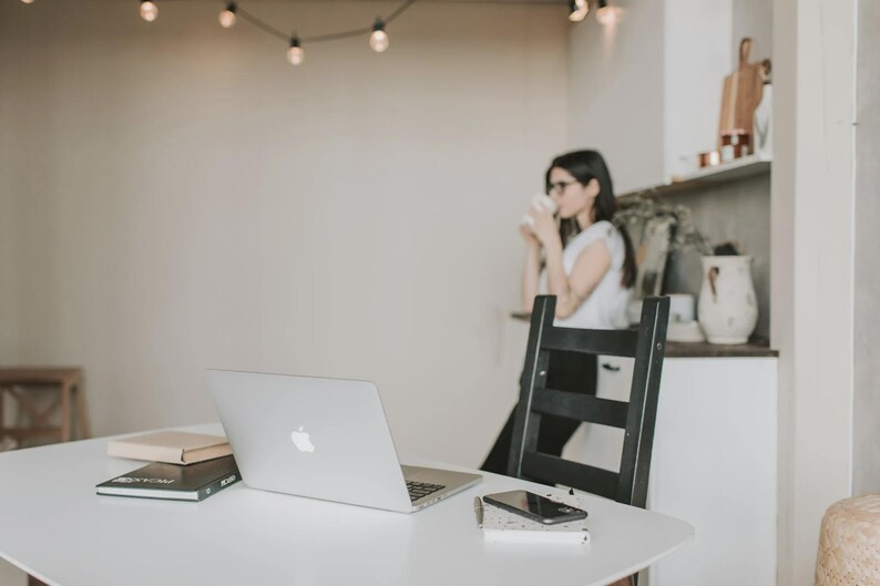 May include: A contemporary workspace with a white table holding a laptop, notebook, and smartphone. A black chair is behind the table. A person is in the background, holding a cup. Overhead, a string of lights adds ambiance.