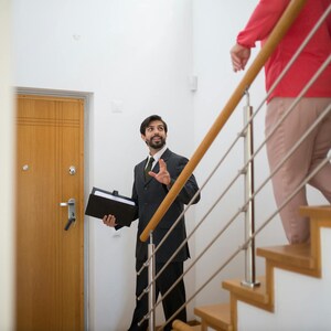 K&ouml;nnte beinhalten: Ein Mann in einem dunklen Anzug und Krawatte gestikuliert, w&auml;hrend er eine schwarze Mappe in der N&auml;he einer Holzt&uuml;r h&auml;lt. Zwei Personen gehen eine Holztreppe mit einem Metallgel&auml;nder hinauf. Eine Person tr&auml;gt ein rotes Hemd und eine beige Hose.
