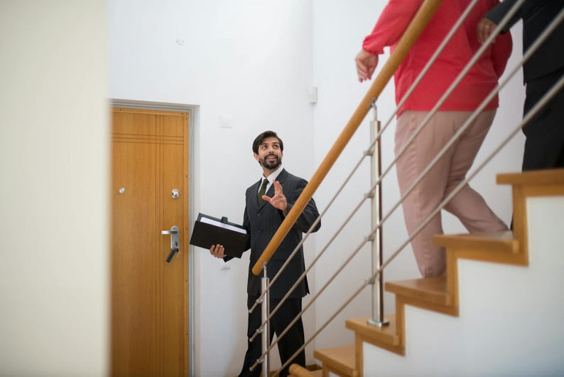 K&ouml;nnte beinhalten: Ein Mann in einem dunklen Anzug und Krawatte gestikuliert, w&auml;hrend er eine schwarze Mappe in der N&auml;he einer Holzt&uuml;r h&auml;lt. Zwei Personen gehen eine Holztreppe mit einem Metallgel&auml;nder hinauf. Eine Person tr&auml;gt ein rotes Hemd und eine beige Hose.