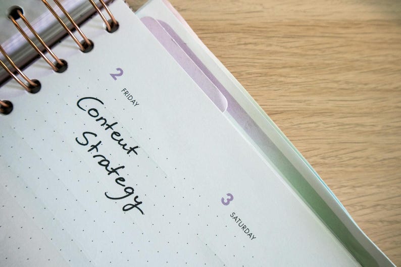 K&ouml;nnte beinhalten: Nahaufnahme eines offenen Spiralnotizbuchs auf einer Holzoberfl&auml;che. Die Seite ist mit Freitag, dem 2. datiert und enth&auml;lt die handschriftlichen Worte "Content Strategy". Das Notizbuch hat einen hellen Einband mit einem gepunkteten Muster.