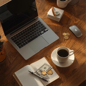 K&ouml;nnte beinhalten: Ein Laptop, eine Kaffeetasse und ein Notizbuch mit Geld und M&uuml;nzen liegen auf einem Holzschreibtisch. Eine kabellose Maus und ein Stapel Notizbl&ouml;cke sind ebenfalls vorhanden. Die Szene deutet auf einen Arbeitsplatz oder ein Homeoffice hin.