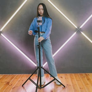 May include: A person in a blue top and jeans adjusts a smartphone on a black tripod. The background features a gray wall with a neon light design. The floor is made of wooden planks.