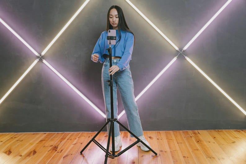 May include: A person in a blue top and jeans adjusts a smartphone on a black tripod. The background features a gray wall with a neon light design. The floor is made of wooden planks.
