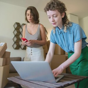 May include: Two people are in a room with cardboard boxes and a laptop. One person is using the laptop, while the other is holding a red smartphone. A lamp and clothing rack are in the background.