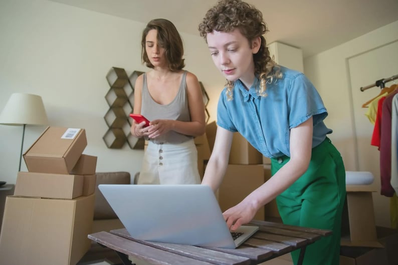 May include: Two people are in a room with cardboard boxes and a laptop. One person is using the laptop, while the other is holding a red smartphone. A lamp and clothing rack are in the background.