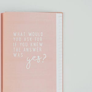 以下が含まれることがあります： 明るいピンク色の表紙の開いたジャーナル。テキスト「WHAT WOULD YOU ASK FOR IF YOU KNEW THE ANSWER WAS yes?」が白で印刷されています。ジャーナルのページには罫線があり、製本が見えます。