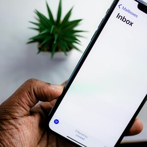 May include: A black smartphone displaying an email inbox labeled "Inbox" on a white screen. The phone is held in a dark-skinned hand. A small green plant is in the background.