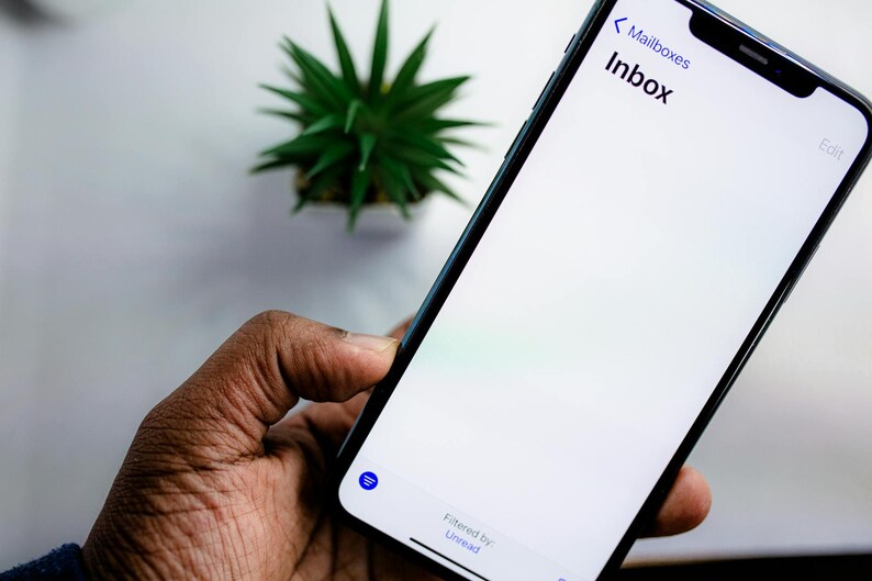 May include: A black smartphone displaying an email inbox labeled "Inbox" on a white screen. The phone is held in a dark-skinned hand. A small green plant is in the background.