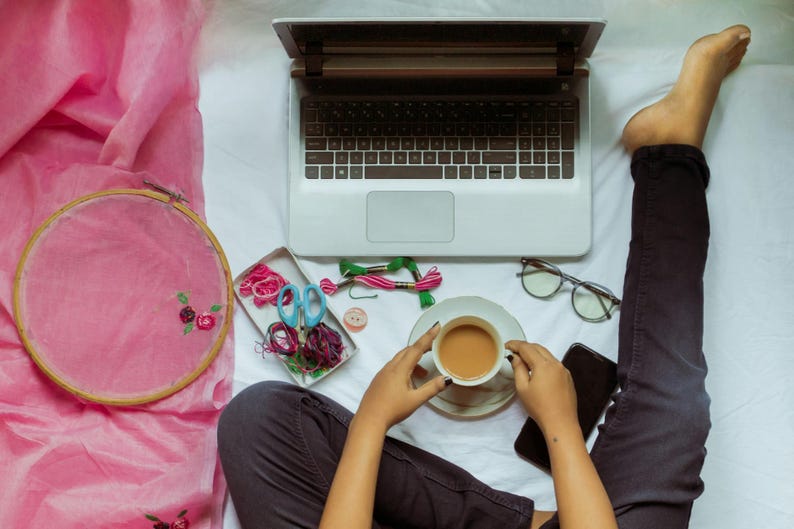 K&ouml;nnte beinhalten: Ein Laptop, ein Stickrahmen und Bastelmaterialien sind auf einer wei&szlig;en Oberfl&auml;che angeordnet. Eine Tasse Kaffee steht in der N&auml;he einer Brille und eines Smartphones. Die Szene deutet auf einen kreativen Arbeitsplatz hin.
