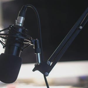 K&ouml;nnte beinhalten: Ein schwarzes Mikrofon mit Schaumstoffabdeckung ist an einem schwarzen, verstellbaren Arm befestigt. Das Mikrofon ist f&uuml;r die Aufnahme von Audio konzipiert. Der Hintergrund ist unscharf, was auf ein Studio oder einen Arbeitsplatz hindeutet.