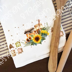 May include: A white kitchen towel with a printed design of honey jars, sunflowers, and beehives. The design is in shades of yellow, brown, and green.