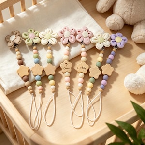 May include: Seven pacifier clips featuring fabric flower attachments in various pastel colors, connected to strings of round silicone beads and a single wooden star-shaped bead. Two of the wooden stars are engraved with the names Emma and Melia.