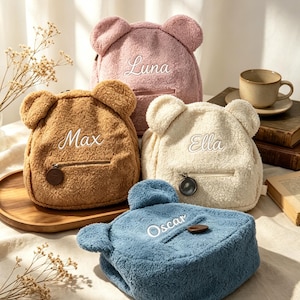 Könnte beinhalten: Vier teddybärförmige Rucksäcke in Rosa, Braun, Creme und Blau. Jeder Rucksack hat gestickte Namen: Luna, Max, Ella und Oscar. Sie haben Bärenohren, einen Reißverschluss und ein kleines Holzknopfdetail.