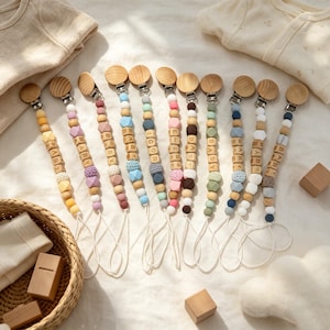 Puede incluir: Una colección de once sujetachupetes dispuestos en fila, cada uno con un clip de madera redondo, una serie de cuentas de madera y silicona de varios colores, algunos con cubos de madera con las letras BABY, IGOR o ELISE.