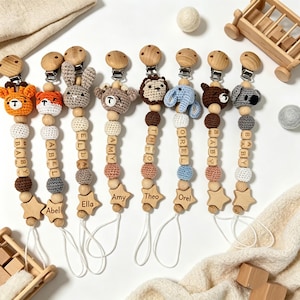 May include: Eight pacifier clips featuring crocheted animal heads including a giraffe, fox, rabbit, bear, lion, elephant, and koala. Each clip has wooden beads, some with letters spelling names like Amy, Theo, and Orel, and a wooden star at the base.