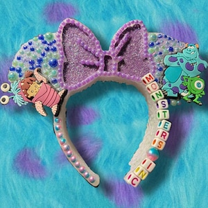 May include: A decorative headband featuring a large, glittery purple bow and character figures from the movie "Monsters, Inc." The headband has the words "Monsters Inc" spelled out in letter beads. The headband is decorated with blue and green beads.
