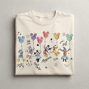 Könnte beinhalten: Ein cremefarbenes T-Shirt mit Rundhalsausschnitt und einem Grafikdruck von Disney-Figuren wie Donald Duck, Daisy Duck, Minnie Maus, Mickey Maus, Pluto und Goofy, die bunte Luftballons halten, ergänzt durch ihre handschriftlichen Signaturen.