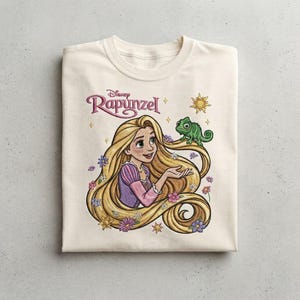 Könnte beinhalten: Ein cremefarbenes T-Shirt mit einem Grafikdruck der Disney-Figur Rapunzel mit langem blonden Haar, einem grünen Chamäleon und kleinen bunten Blumen. Der Text Disney Rapunzel ist in einer stilisierten violetten Schrift über der Figur gedruckt.