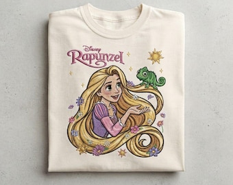 May include: A cream-colored t-shirt featuring a graphic print of the Disney character Rapunzel with long blonde hair, a green chameleon, and small colorful flowers. The text Disney Rapunzel is printed in a stylized purple font above the character.