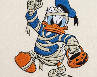 May include: An embroidered patch featuring a cartoon duck dressed as a mummy, wrapped in beige bandages over a blue shirt. The duck wears a blue cap, has an orange beak and feet, and holds a small orange jack-o'-lantern bucket with a black face.