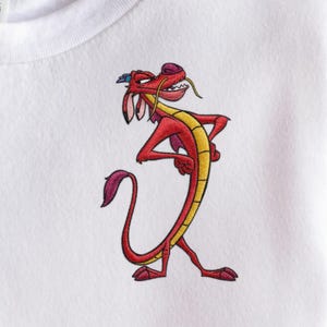 May include: White t-shirt featuring an embroidered red dragon with yellow accents. The dragon has a confident pose with its arms crossed and a stern expression. The design is centered on the shirt.