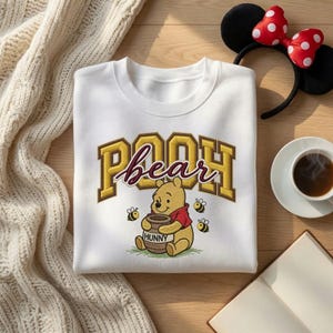 May include: A white crewneck sweatshirt featuring a graphic of Winnie the Pooh holding a brown honey pot labelled HUNNY. The text POOH is printed in large yellow block letters with a dark outline, and the word bear is written in maroon script.