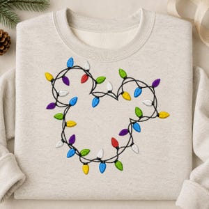 May include: A sand-colored crewneck sweatshirt featuring an embroidered design of a black wire string of multi-colored Christmas lights arranged in the shape of a mouse head silhouette, with red, blue, green, yellow, purple, and white bulbs.