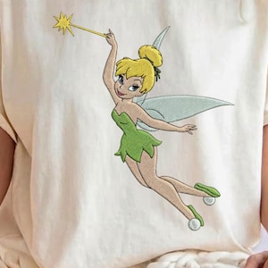 Può includere: Una t-shirt color crema con il ricamo di una fata con i capelli biondi raccolti in uno chignon, che indossa un vestito verde e scarpe con pon-pon bianchi. La fata ha ali traslucide e tiene una bacchetta magica con una stella gialla.