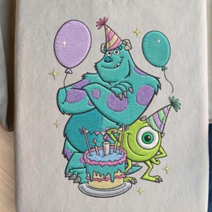 May include: Embroidered design featuring the characters Sully and Mike from Monsters, Inc. Sully, in teal with purple spots, wears a party hat and holds a purple balloon. Mike, in green, also wears a party hat. A birthday cake with candles is in front.