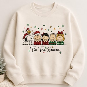 以下が含まれることがあります： クリーム色のクルーネックのスウェットシャツ。サンタ帽をかぶり、クリスマスの装いをしたスヌーピー、ウッドストック、ピーナッツの仲間たちが刺繍されています。キャラクターの下には、黒い筆記体で「Tis The Season」という文字が記されています。