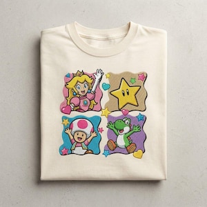 Puede incluir: Una camiseta de color crema con un estampado gráfico de cuatro paneles cuadrados. Los paneles muestran a la Princesa Peach, una estrella amarilla, Toad y Yoshi. El diseño está decorado con pequeñas estrellas y corazones de colores.