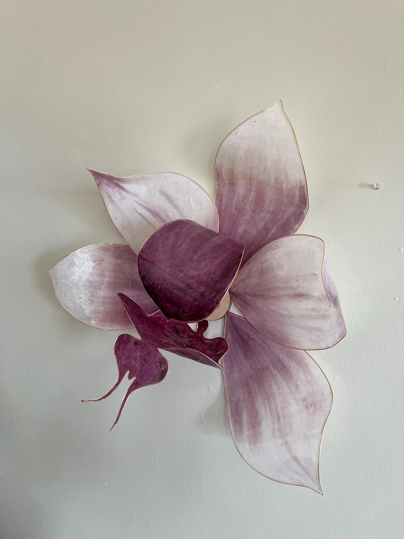 May include: A wall-mounted flower sculpture featuring layered, translucent petals in shades of pale pink and deep plum. The petals have thin, metallic-toned edges, and the center displays a dark, saturated purple hue with a distinct, elongated shape.