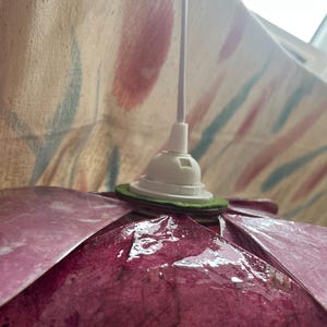 May include: A pendant lamp shade featuring overlapping dark magenta, textured, fibrous petals arranged in a circular pattern. The shade is attached to a white plastic socket and a white electrical cord, with a green felt ring spacer.
